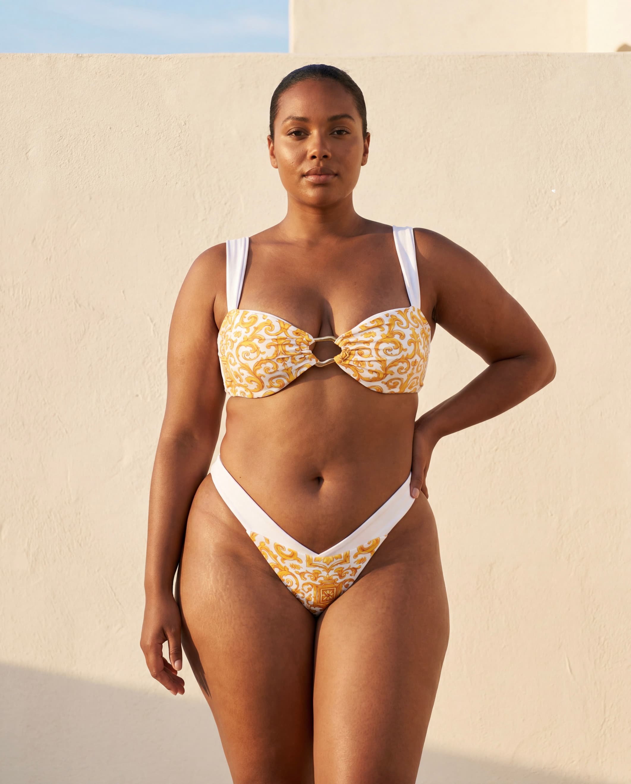  Mujer curvy en bikini Capri Goddess blanco con estampado de mayólica mediterránea dorada y top con aro central, frente a un muro encalado, colección Riviera de Leonika Swimwear