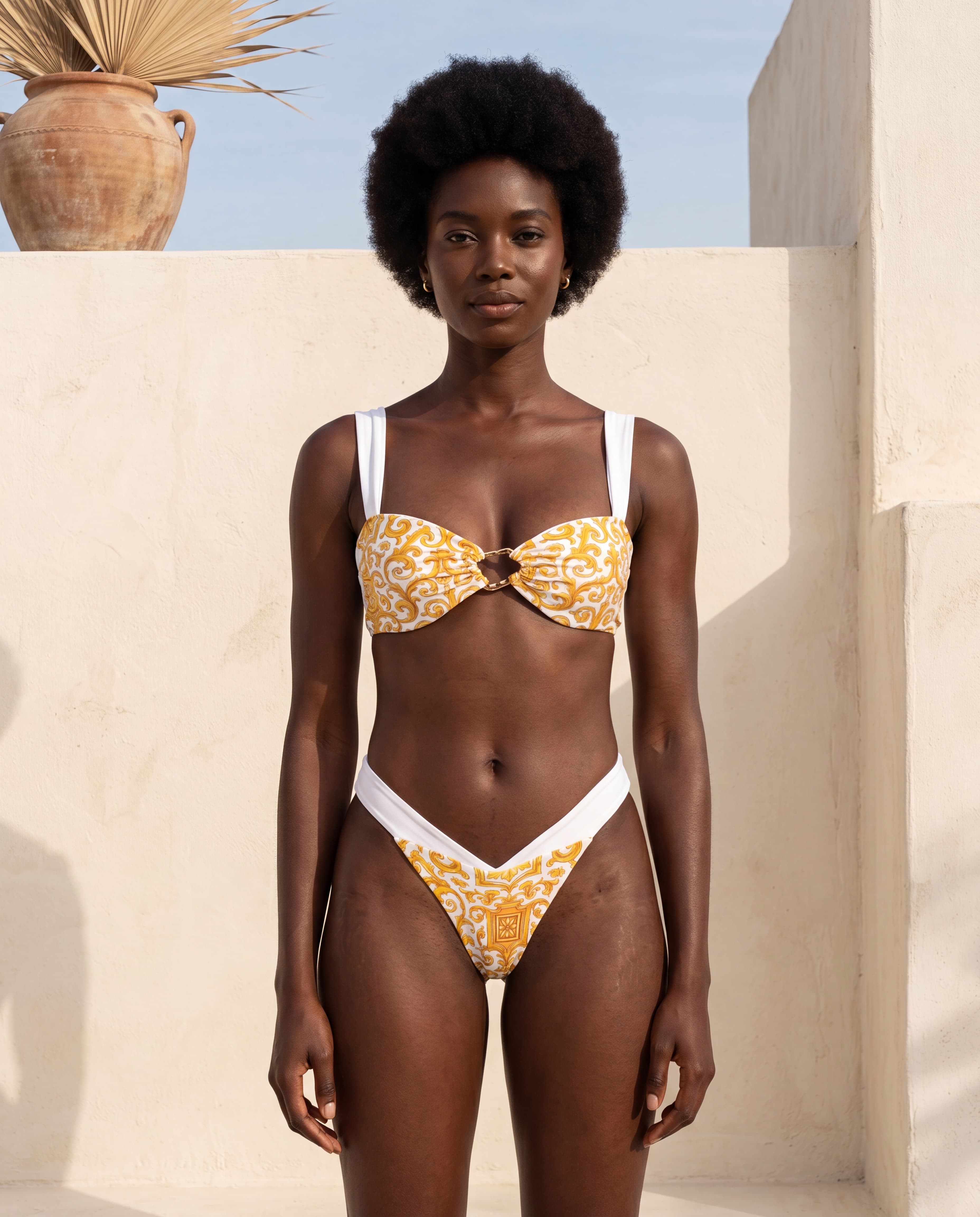Mujer en bikini Capri Goddess blanco con estampado de mayólica mediterránea dorada, en una terraza encalada con una tinaja de barro, colección Riviera de Leonika Swimwear