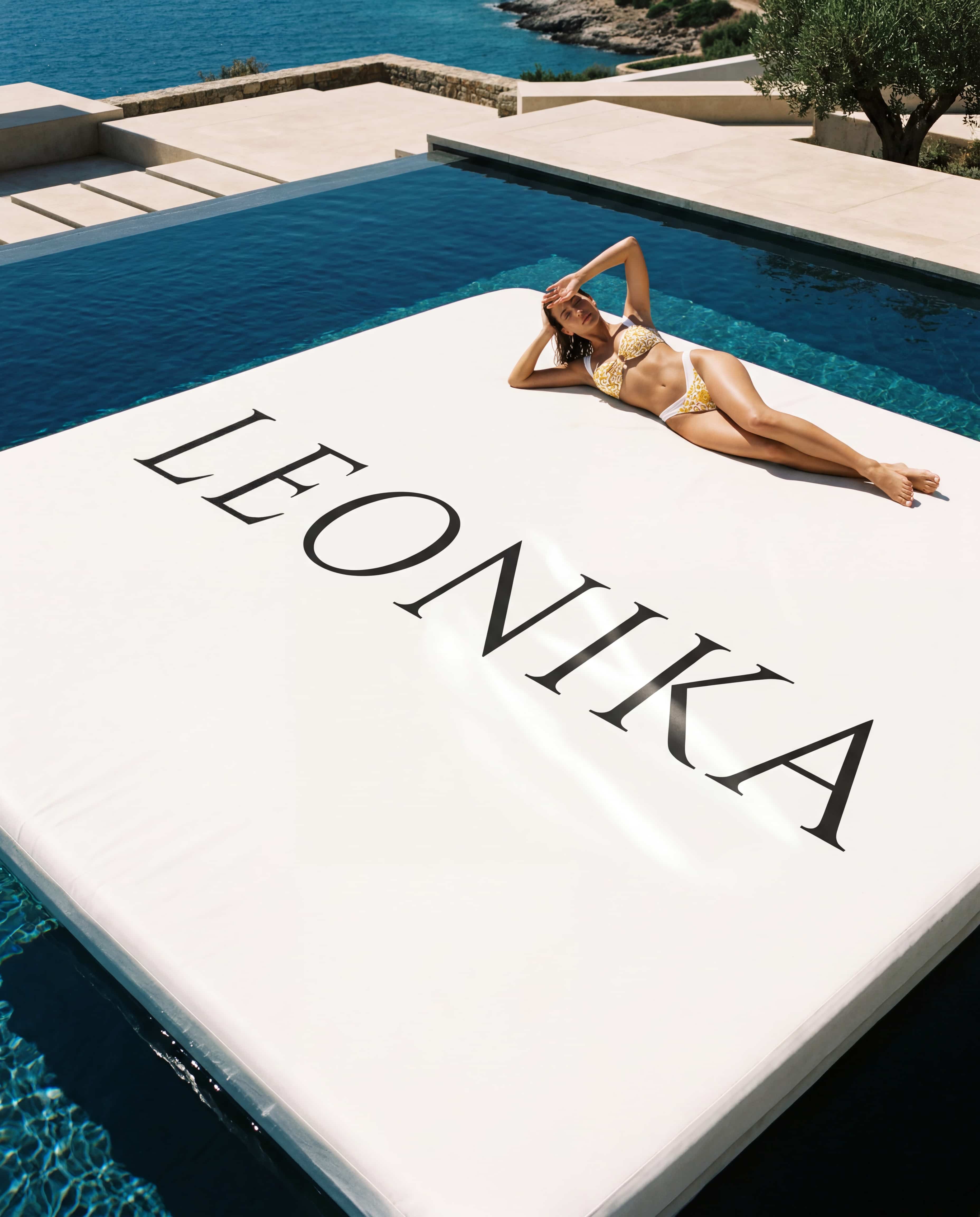 Mujer en bikini Capri Goddess blanco y dorado recostada junto a una piscina infinita mediterránea, con el logotipo de Leonika, imagen de marca de la colección Riviera Resort